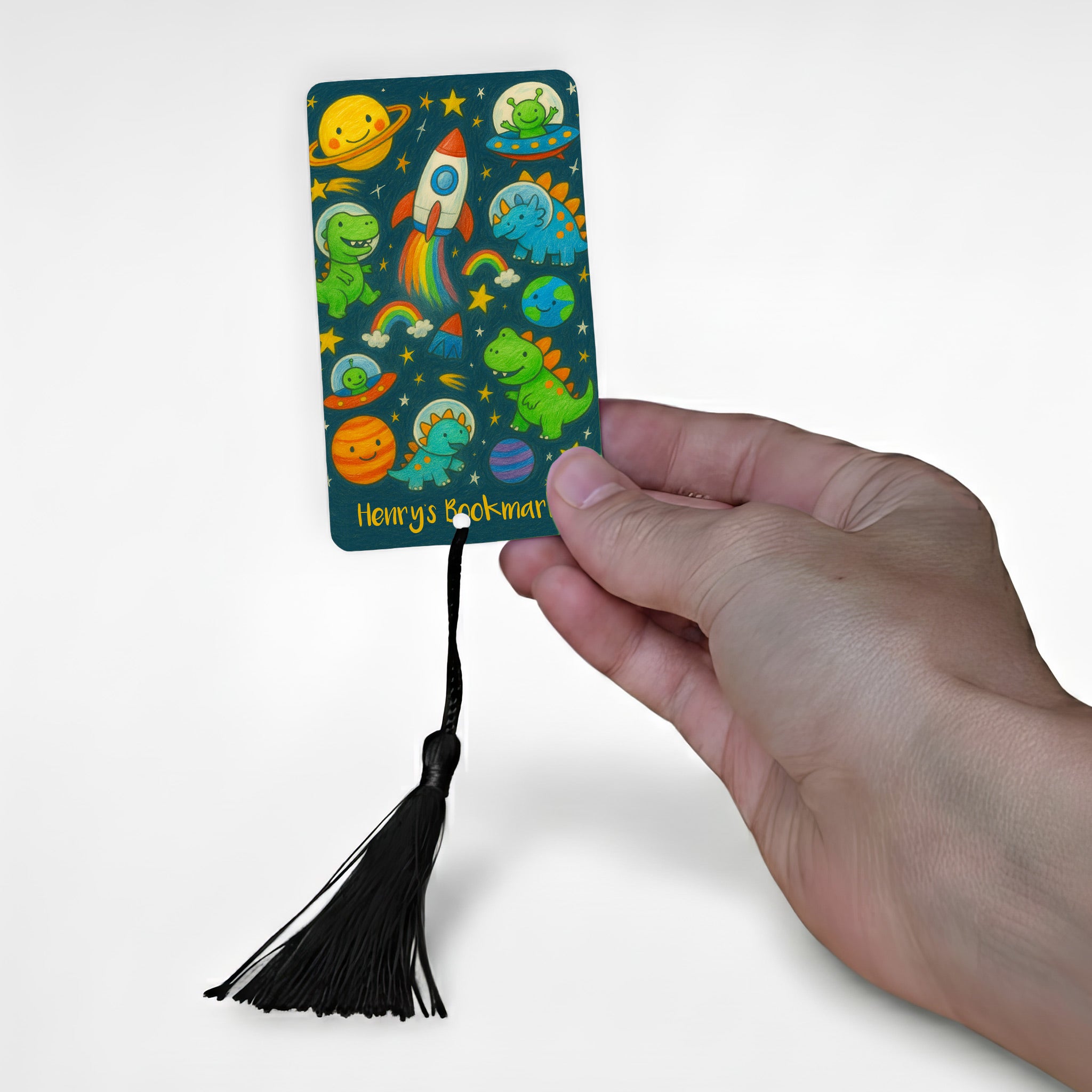 Personalised children’s metal bookmark with colourful space dinosaurs, planets and rocket ship design, shown held in hand with a black tassel – custom kids space bookmark handmade in the UK.