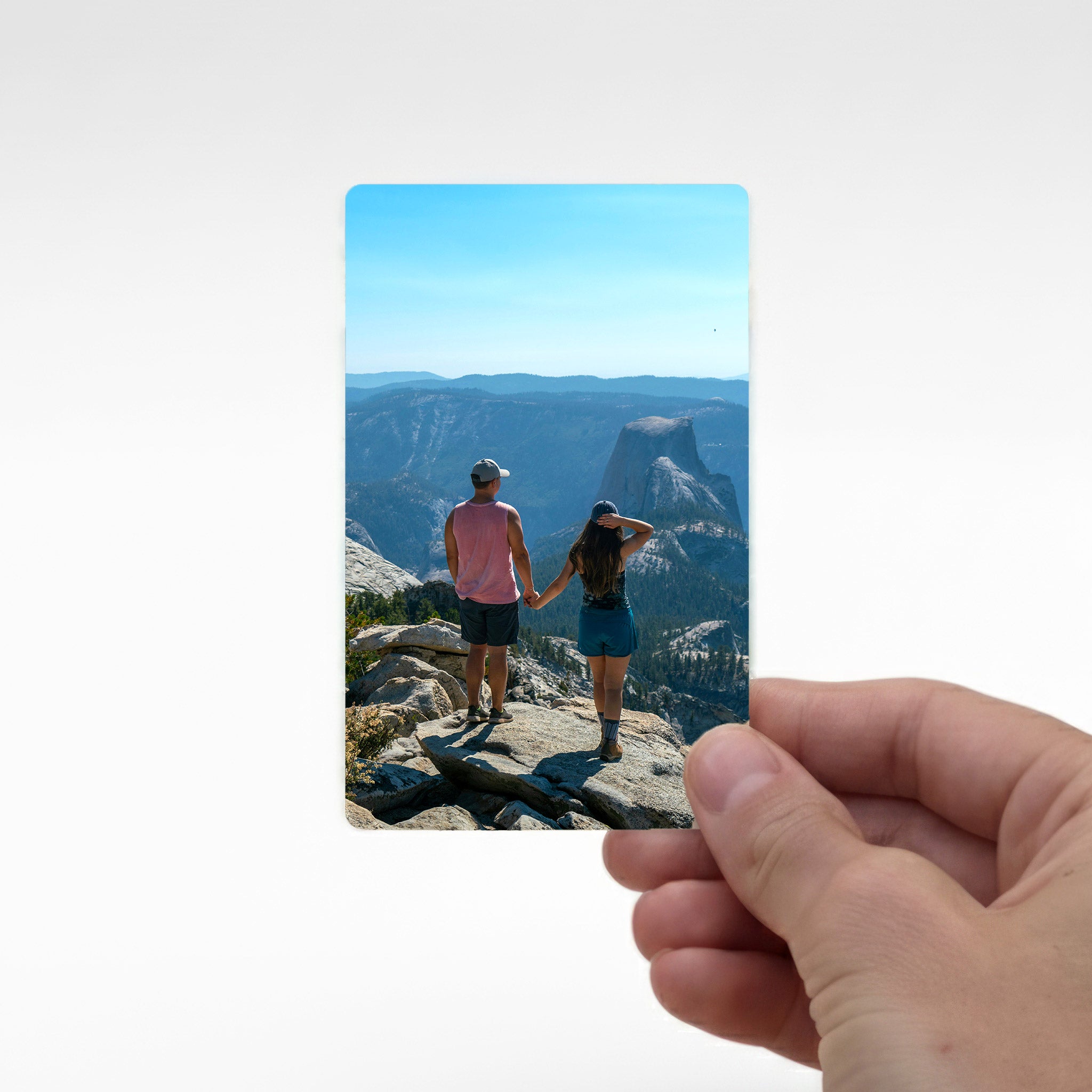Hand holding a personalised metal photo wallet card printed with a couple standing on a mountain viewpoint, demonstrating wallet-size fit. Great for preserving travel memories.