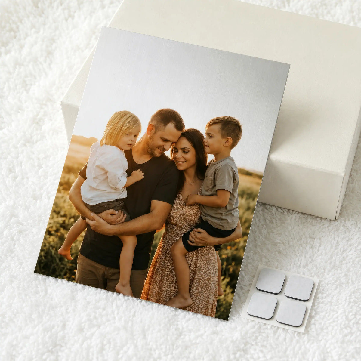 A custom printed a4 size metal photo of a family of four displayed on a white textured blanket next to a white gift box and four silver adhesive velcro patches.