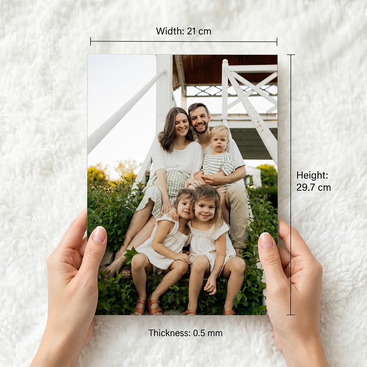 A person's hands hold a custom metal a4 size photo print showing a family of five, with dimension lines and text indicating the print's size: 21 cm width, 29.7 cm height, and 0.5 mm thickness.